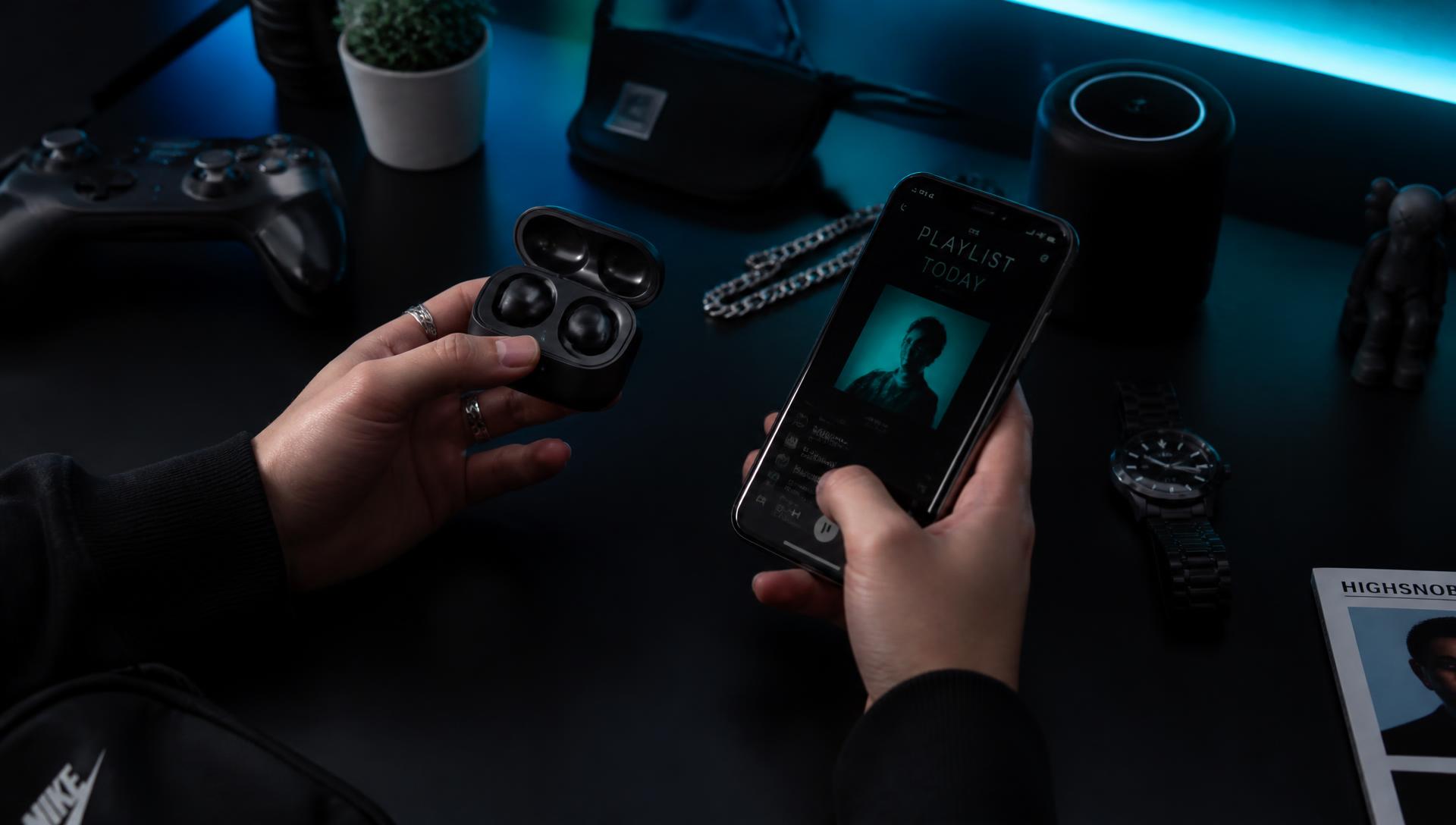 Young person holding wireless earbuds and smartphone on a dark desk with cyan accent lighting